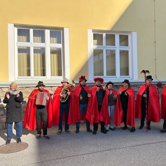 Eine Gruppe von Menschen in roten Umhängen und Hüten, einschließlich einer Frau in einem schwarzen Mantel, posiert für ein Foto vor einem Gebäude mit gelben Wänden und weißen Fenstern.