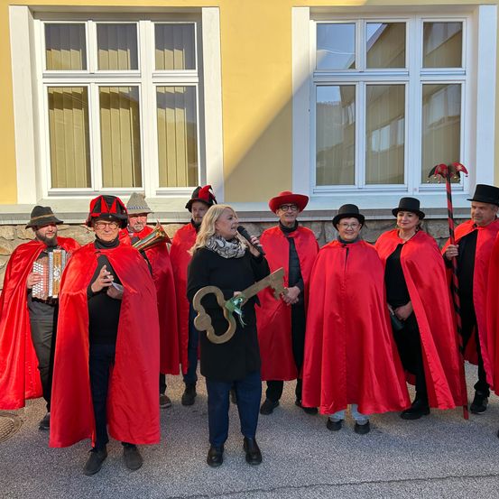 Eine Gruppe von Menschen in roten Kostümen mit Hüten und Umhängen steht vor einem Gebäude. Eine Frau in der Mitte hält ein Mikrofon und einen Schlüssel. Die Gruppe umfasst Musiker, die Instrumente spielen.