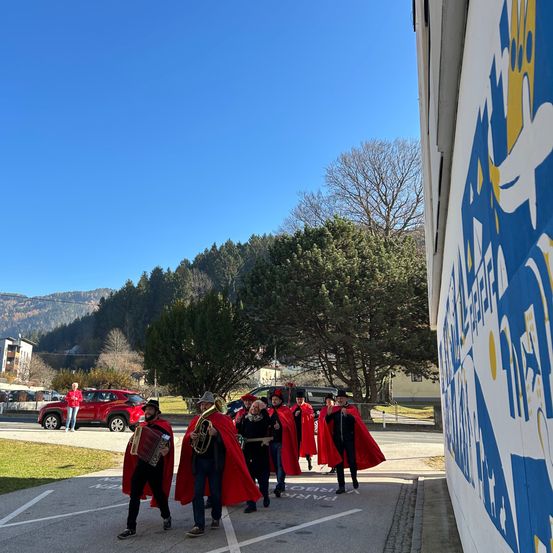 Eine Gruppe von Musikern in roten Umhängen geht auf einer Straße und spielt Instrumente. Hinter ihnen steht ein rotes Auto und eine Person. Im Hintergrund sind Bäume und Berge zu sehen.
