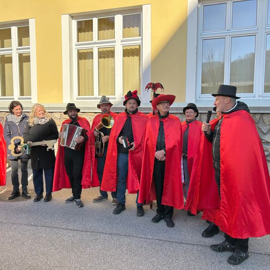 Eine Gruppe von Menschen in roten Umhängen und Hüten steht vor einem Gebäude, ein Mann mit einem Mikrofon und ein anderer mit einem Schlüssel und Akkordeon.