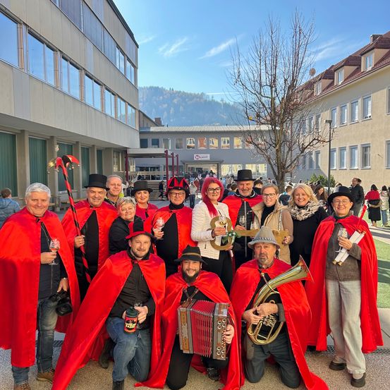 Eine Gruppe von Menschen in roten Umhängen und Hüten posiert für ein Foto vor einem Gebäude, wobei einer einen Tuba und der andere eine Flasche hält.