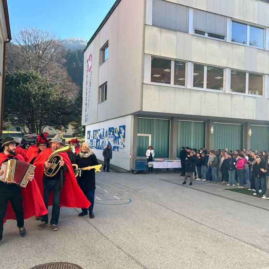 Eine Gruppe Musiker in roten Roben und Hüten tritt vor einer Menschenmenge vor einem Schulgebäude auf.