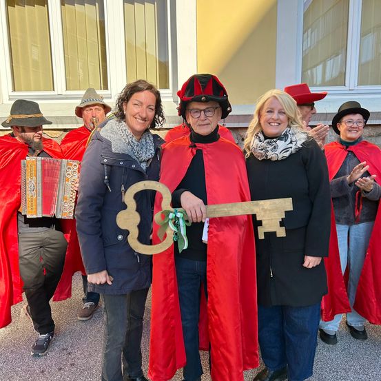 Eine Gruppe von Erwachsenen in roten Umhängen und Hüten steht vor einem Gebäude. Zwei Frauen und ein Mann in einem roten Umhang halten einen großen Holzschlüssel. Hinter ihnen spielen Menschen Akkordeons.