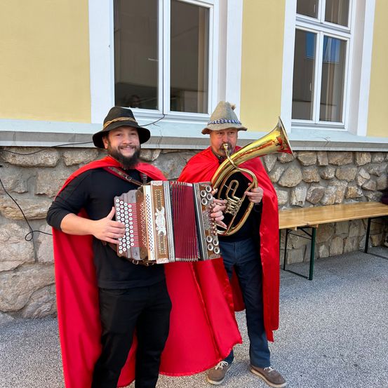 Zwei Männer in roten Umhängen stehen draußen, einer spielt Akkordeon und der andere Posaune, nahe einer Steinmauer und einem Holztisch.