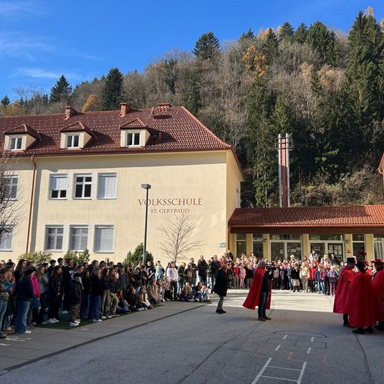 Eine Menschenmenge versammelt sich vor einem Gebäude mit rotem Dach. Einige Personen tragen rote Umhänge und Hüte. Das Gebäude trägt den Namen 'VOLKSSCHULE ST. GERTRAUD'. Im Hintergrund befinden sich Bäume und ein Turm.
