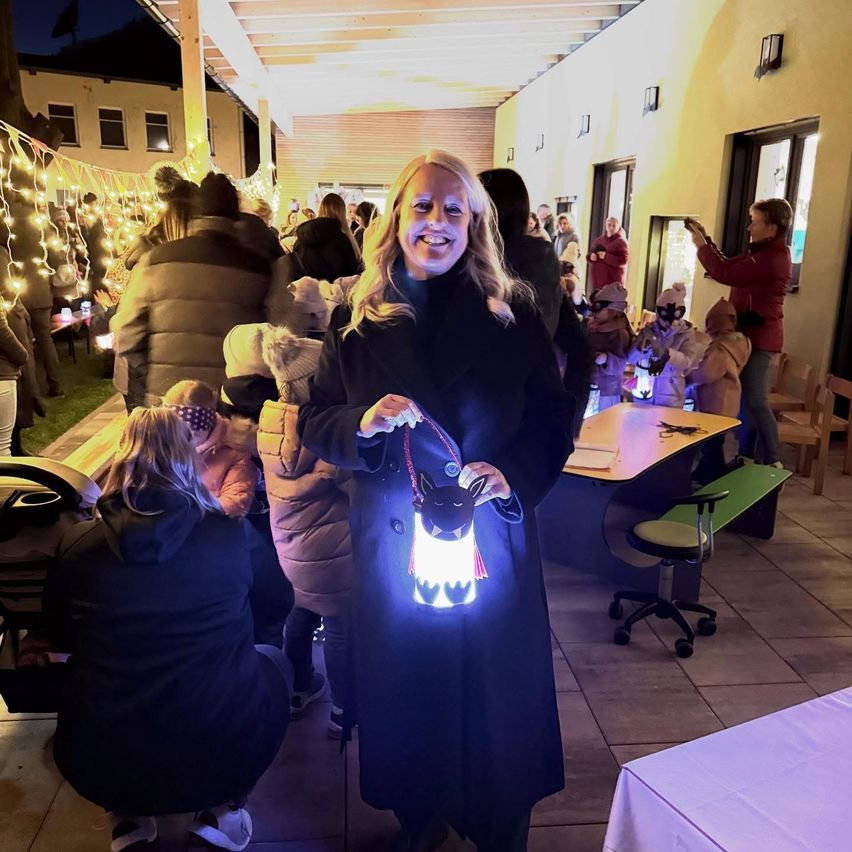 Eine lächelnde Frau hält eine Laterne bei einer Veranstaltung im Freien, umgeben von Kindern und Erwachsenen, beleuchtet von Lichterketten und warmen Lichtern.