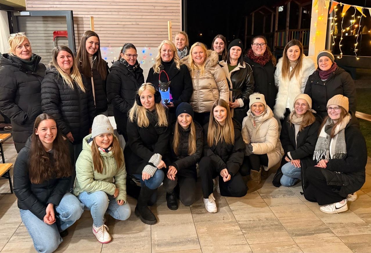 Eine Gruppe von Frauen in Winterkleidung posiert für ein Foto, lächelt und hält eine Geschenktüte. Sie sind drinnen mit einer Holzwand und bunten Lichtern.