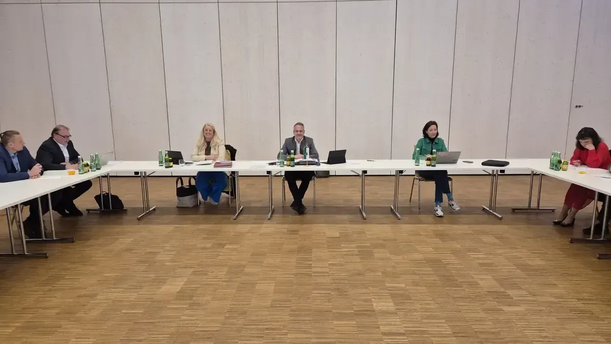 Vier Personen sitzen an einem langen Tisch mit Laptops. Flaschen und andere Gegenstände stehen auf dem Tisch. Der Raum hat eine weiße Wand und einen Holzboden.