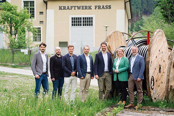Sieben Personen posieren für ein Foto vor Kraftwerk Frass, mit drei großen Holzspulen dahinter. Bäume und Gras sind um sie herum.