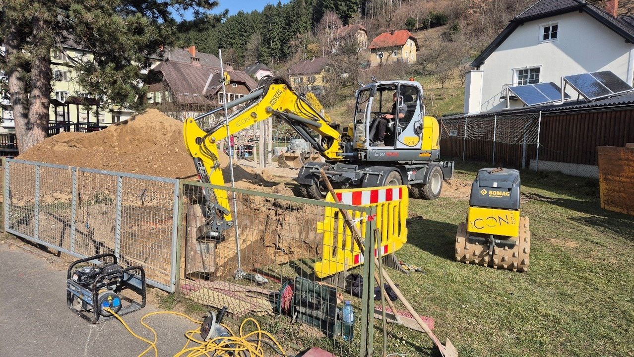 Ein gelber Bagger steht neben einem Zaun auf dem Rasen, mit einem Mann im Inneren. Dahinter befindet sich ein Haufen Erde. In der Nähe sind verschiedene Bauwerkzeuge und Ausrüstungsgegenstände.