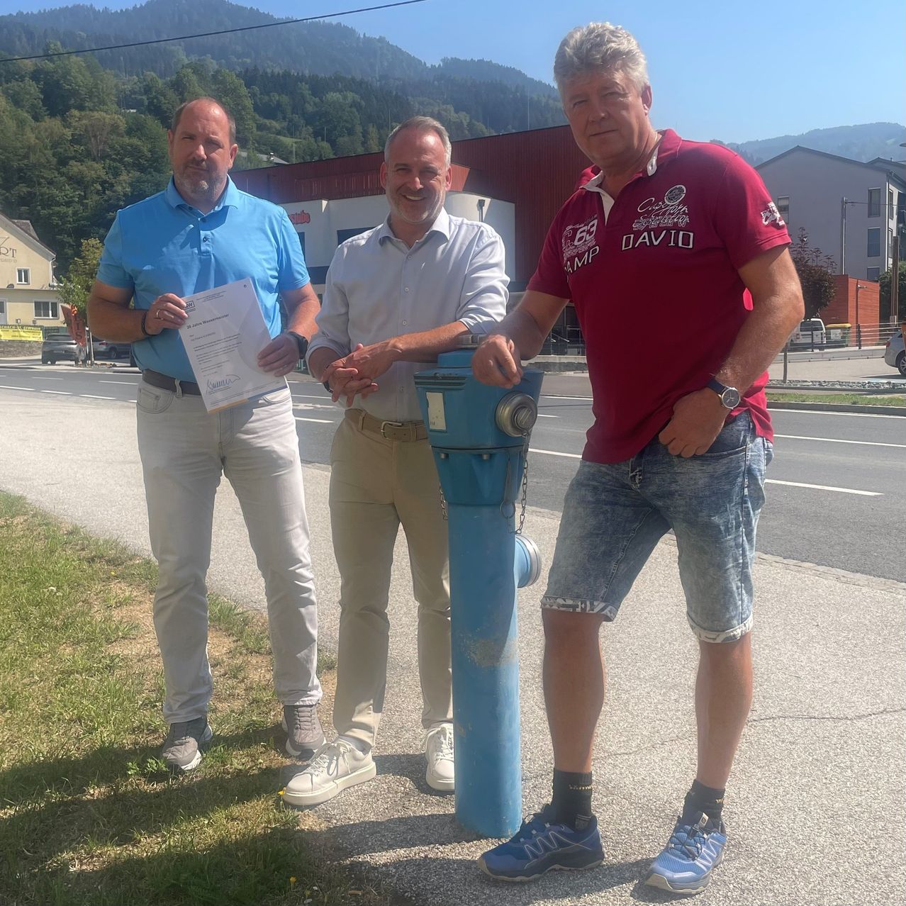 Drei Männer stehen auf einem Bürgersteig, einer hält ein Papier, ein anderer einen blauen Wasserhahn und der dritte trägt ein rotes Shirt. Sie lächeln und scheinen für ein Foto zu posieren. Hinter ihnen ist ein Grasbereich, eine Straße und Gebäude.