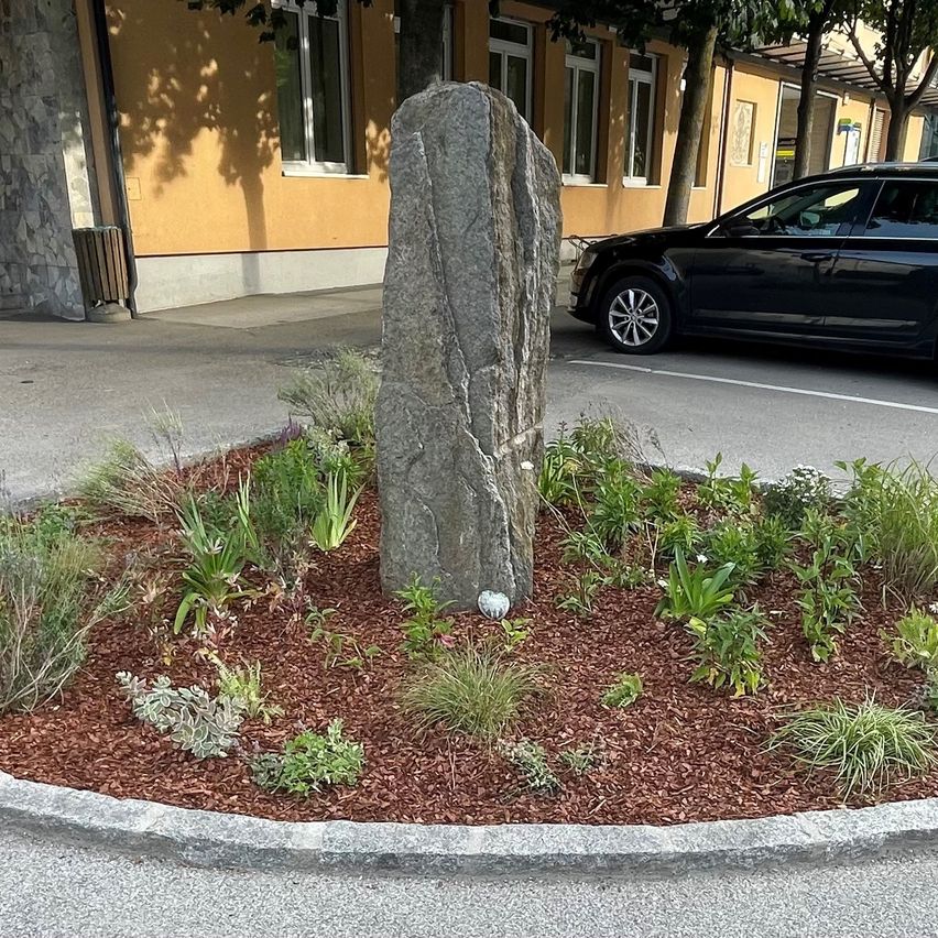 A large stone is in the center of a circular garden bed with red mulch. A black car is parked on the road behind a yellow building.