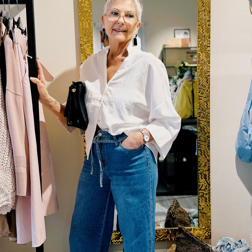 An older woman with glasses and a white blouse stands in a clothing store, holding a black handbag. Behind her, clothes hang on a rack, and a mirror reflects the room.