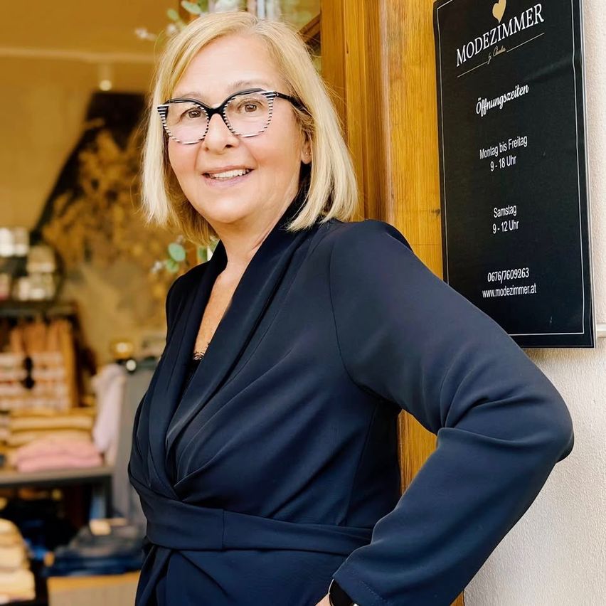 Eine lächelnde Frau mit kurzen blonden Haaren, Brille und dunklem blauem Outfit steht vor einem Ladenschild mit Öffnungszeiten.