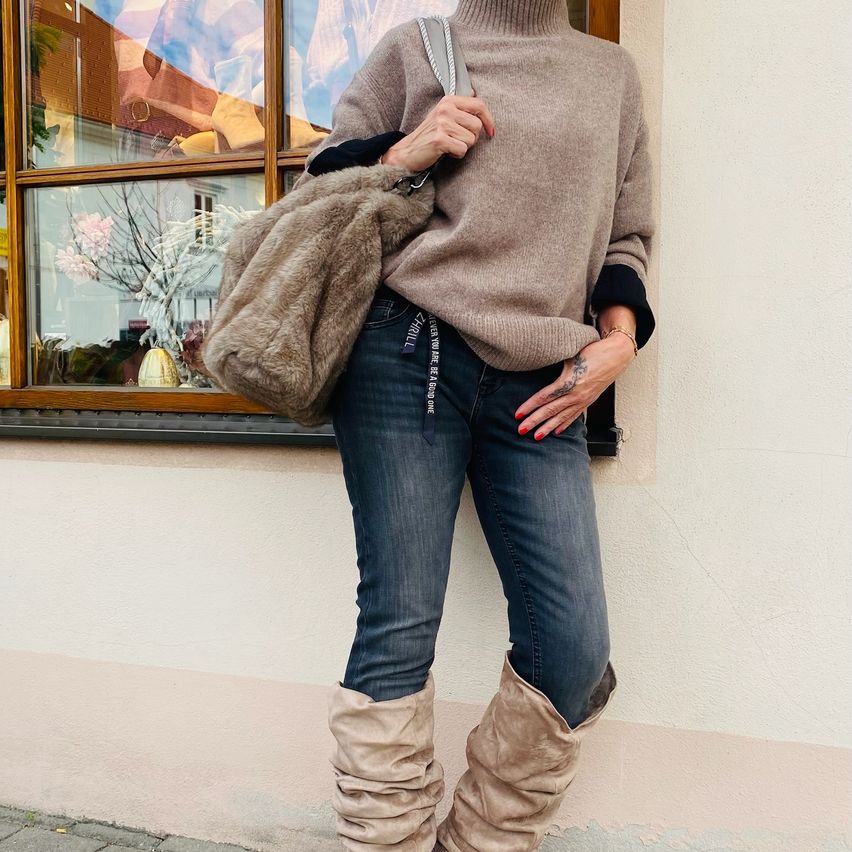A woman stands outside, leaning against a wall, wearing a beige turtleneck, jeans, and beige boots. She holds a fur bag.