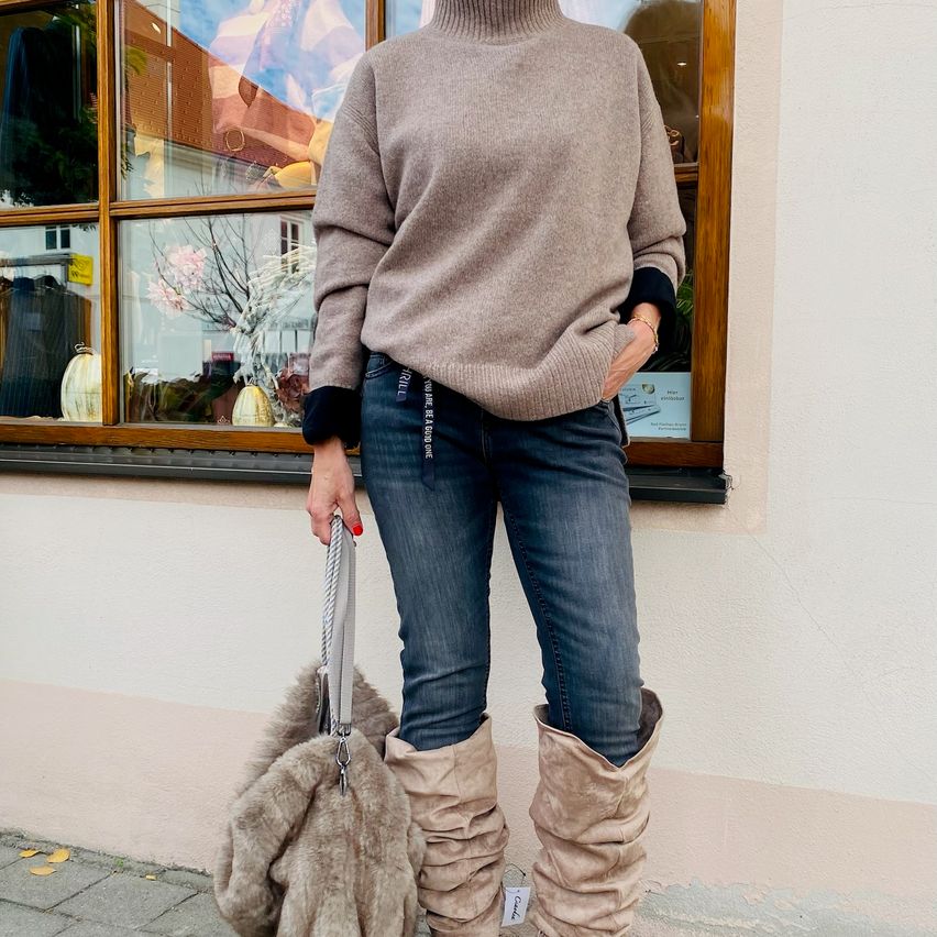A woman wearing a beige turtleneck sweater, blue jeans, and over-the-knee boots stands outside a shop.