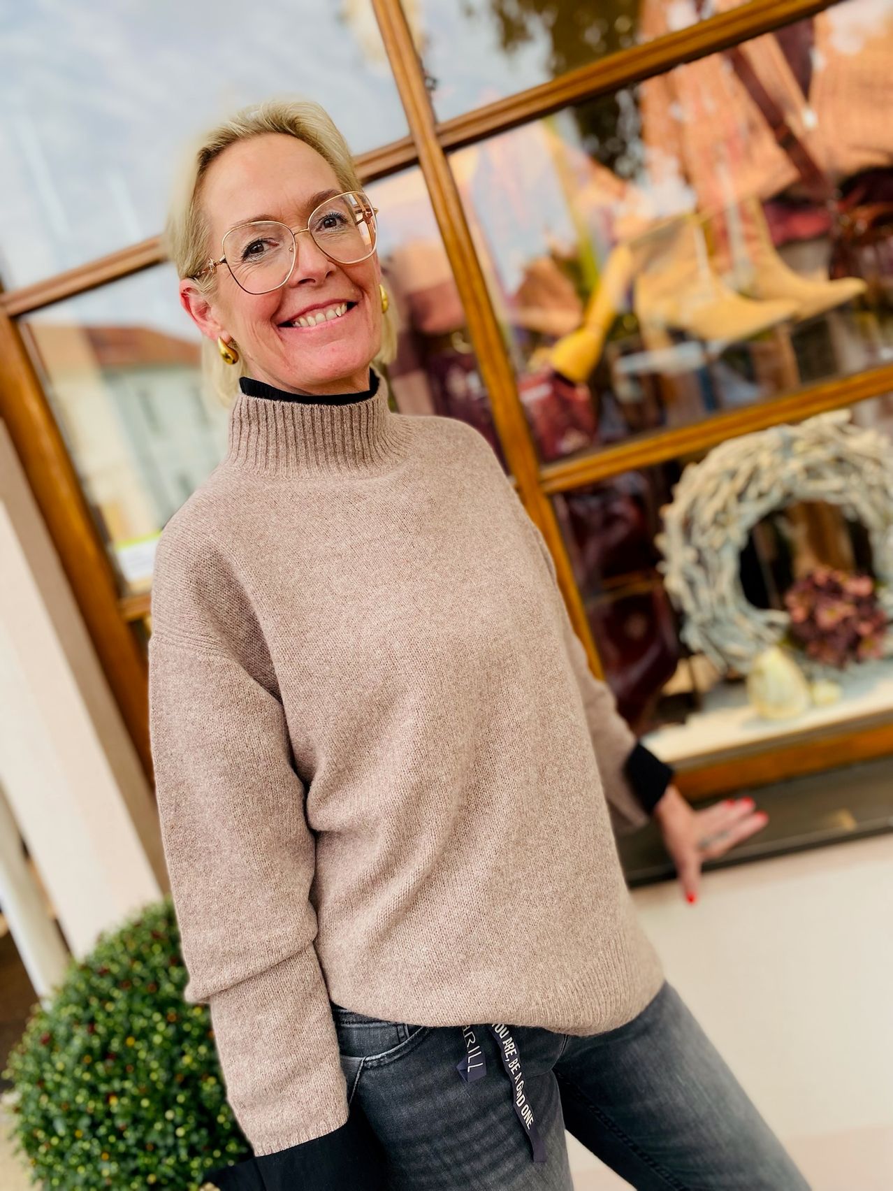 A woman with blonde hair and glasses stands in front of a store window, smiling. She wears a beige turtleneck sweater and jeans. Behind her, the store displays various items, including a wreath and shoes.