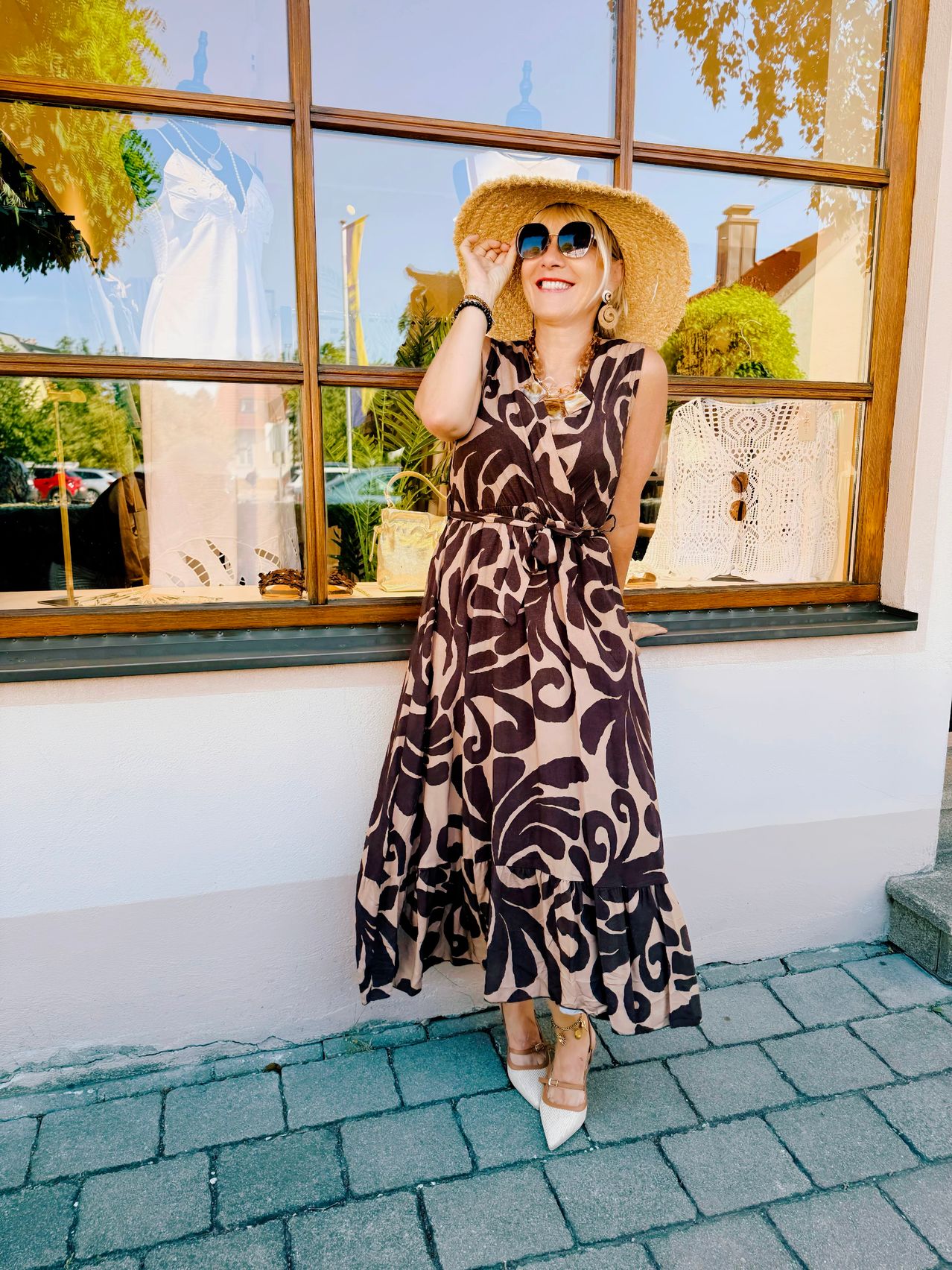 Eine Frau in einem gemusterten Kleid und Hut steht vor einem Boutique, lächelt und posiert für ein Foto. Das Schaufenster zeigt verschiedene Artikel, darunter ein weißes Spitzenkleid.