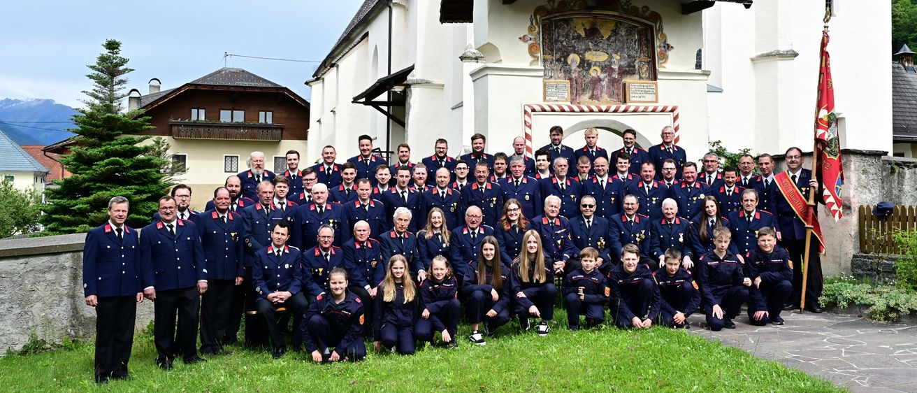 Eine Gruppe uniformierter Personen posiert vor einer Kapelle mit einem verzierten Torbogen. Einige sitzen, andere stehen auf dem Rasen.