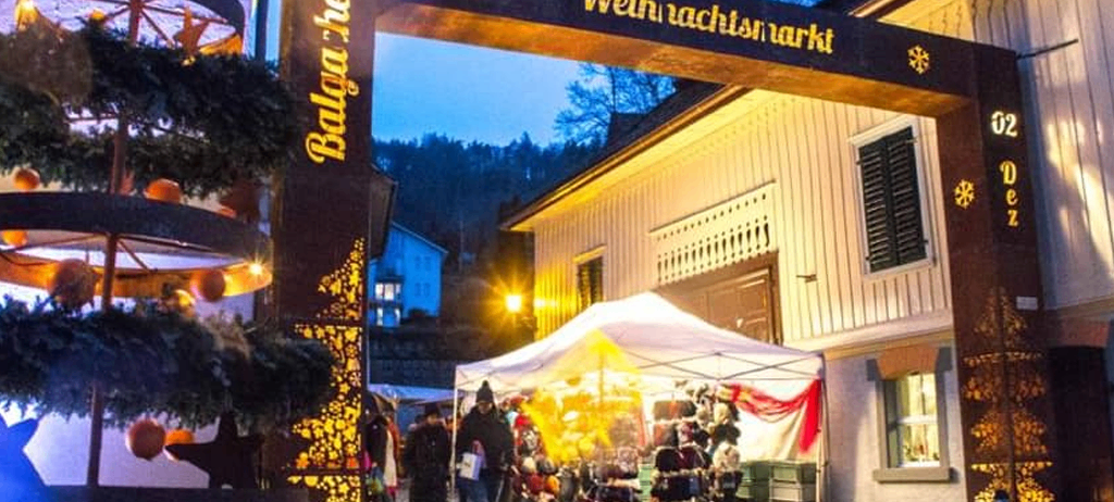 Der Eingang des Balvaracher Weihnachtsmarktes bei Nacht. Der Markt ist mit Weihnachtsdekorationen und Lichtern geschmückt. Menschen gehen herum, und ein Zelt steht in der Mitte.