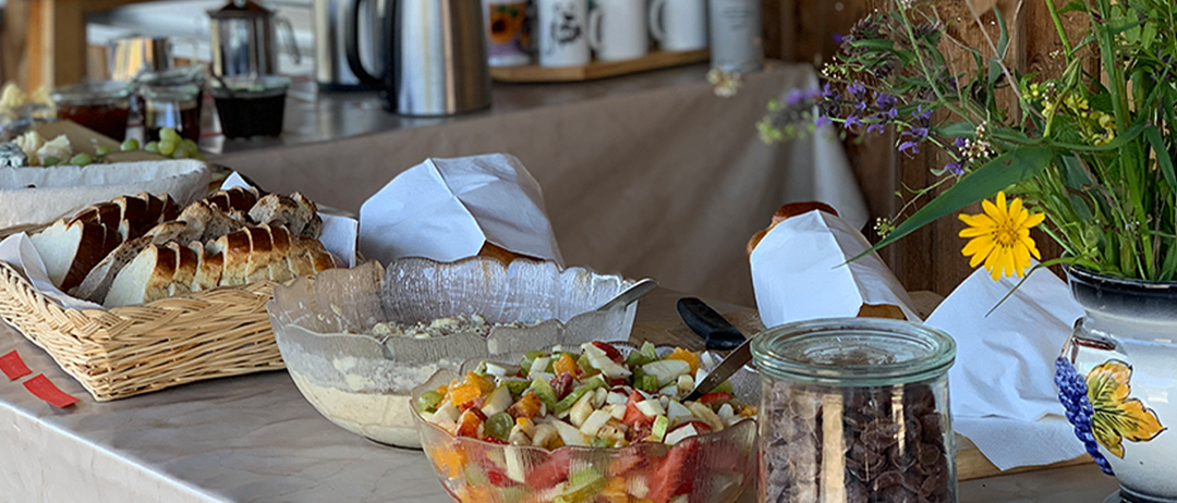 Ein Frühstücksbuffet mit verschiedenen Gerichten, einschließlich einer Obstsalat, Brot und Gebäck. Ein hölzerner Tresen hat einen Wasserkocher, Tassen und Gläser. Ein Blumentopf und ein hölzerne Schild sind im Hintergrund.