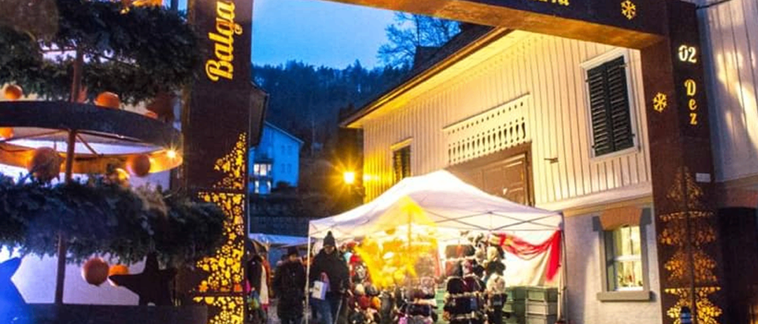 Ein Weihnachtsmarkt mit einem beleuchteten Vordach und Ständen, die verschiedene Artikel verkaufen, umgeben von festlichen Dekorationen und einem Schild, das Weihnachtsmarkt bedeutet.