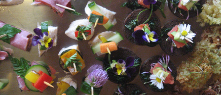 Ein Nahaufnahme von verschiedenen Fingerfoods, die mit essbaren Blumen und Gemüse garniert sind, einschließlich geräuchertes Fleisch, Sandwiches und Gurkenscheiben.