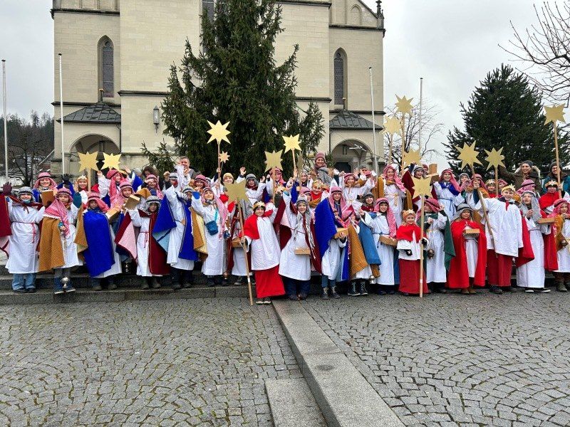 Eine Gruppe von Kindern, die als biblische Figuren gekleidet sind, posiert für ein Foto vor einer Kirche. Sie halten sternförmige Dekorationen und haben festliche Kostüme.