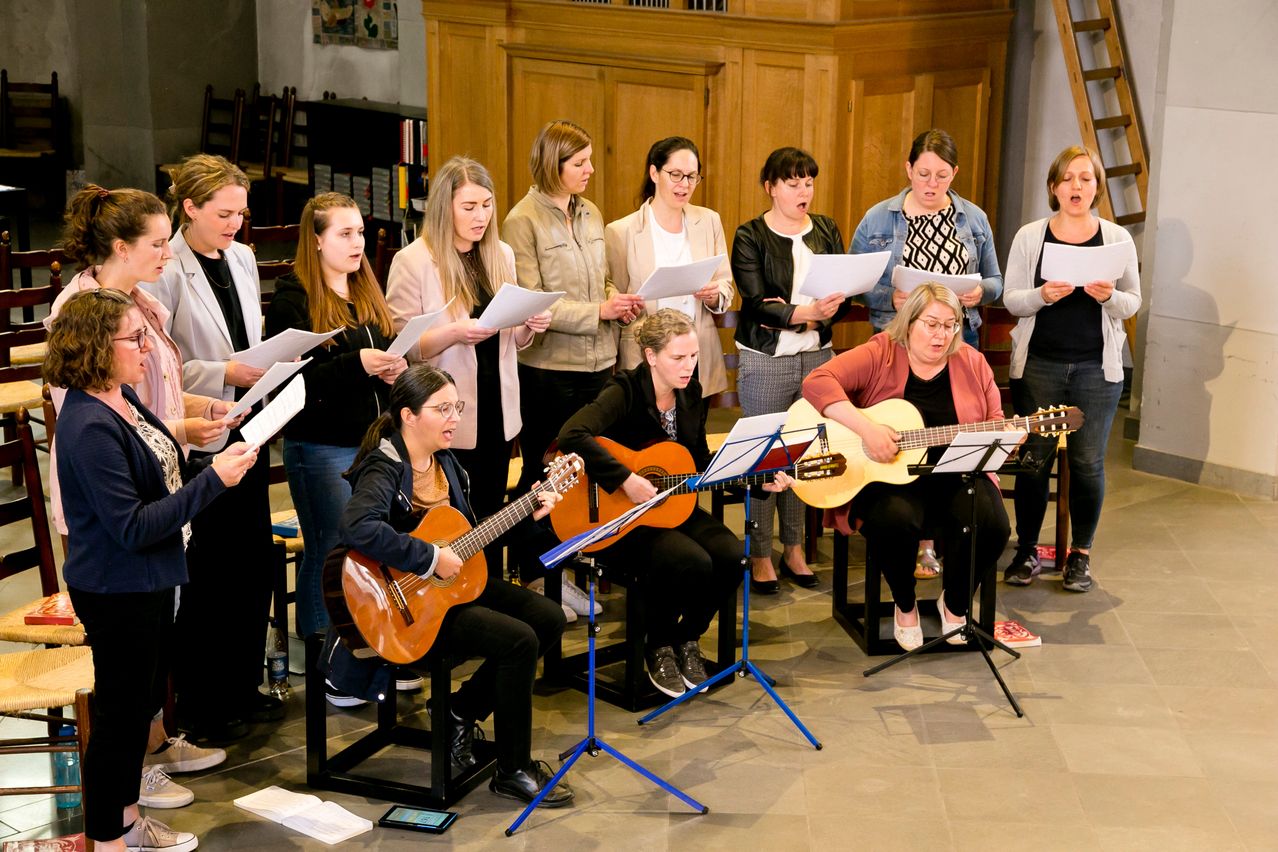Eine Gruppe von Frauen singt in einem Raum, während sie Papiere in der Hand halten. Einige sitzen auf Hockern und spielen Gitarre. Hinter ihnen stehen andere Frauen mit Papieren in der Hand. Die Umgebung ähnelt einer Kirche.