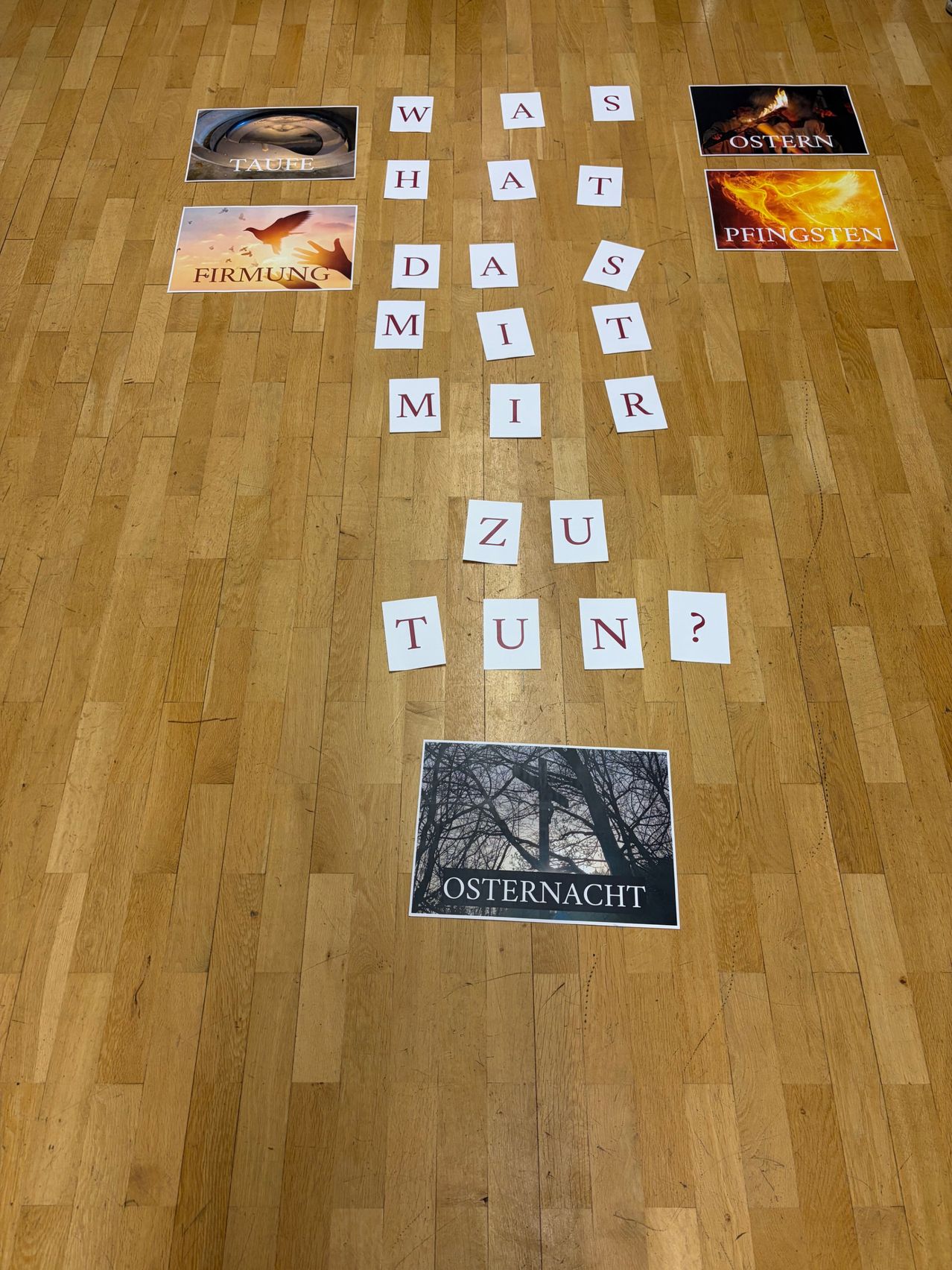 Ein Bild von Buchstaben und Bildern auf einem Holzboden. Buchstaben bilden 'zu tun?' mit Bildern eines Baumes und eines Vogels. Andere Bilder zeigen 'Firmung', 'Taufe', 'Pfingsten' und 'Ostern'.
