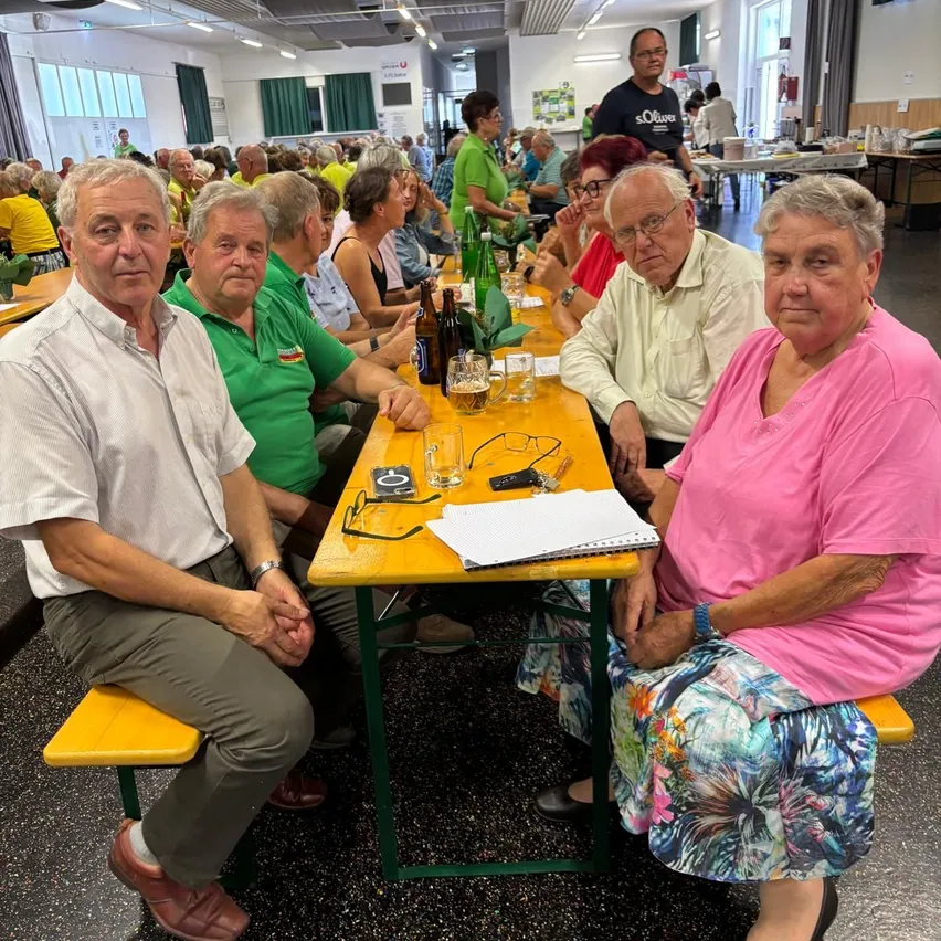 Eine Gruppe älterer Menschen sitzt an einem langen Tisch in einem Saal. Auf dem Tisch stehen Gläser, Flaschen und Notizbücher. Die Menschen tragen Freizeitkleidung. Einige tragen Brillen. Im Hintergrund sind andere Menschen zu sehen. Es gibt Tische und Stühle im Saal. An der Wand hängen Vorhänge.