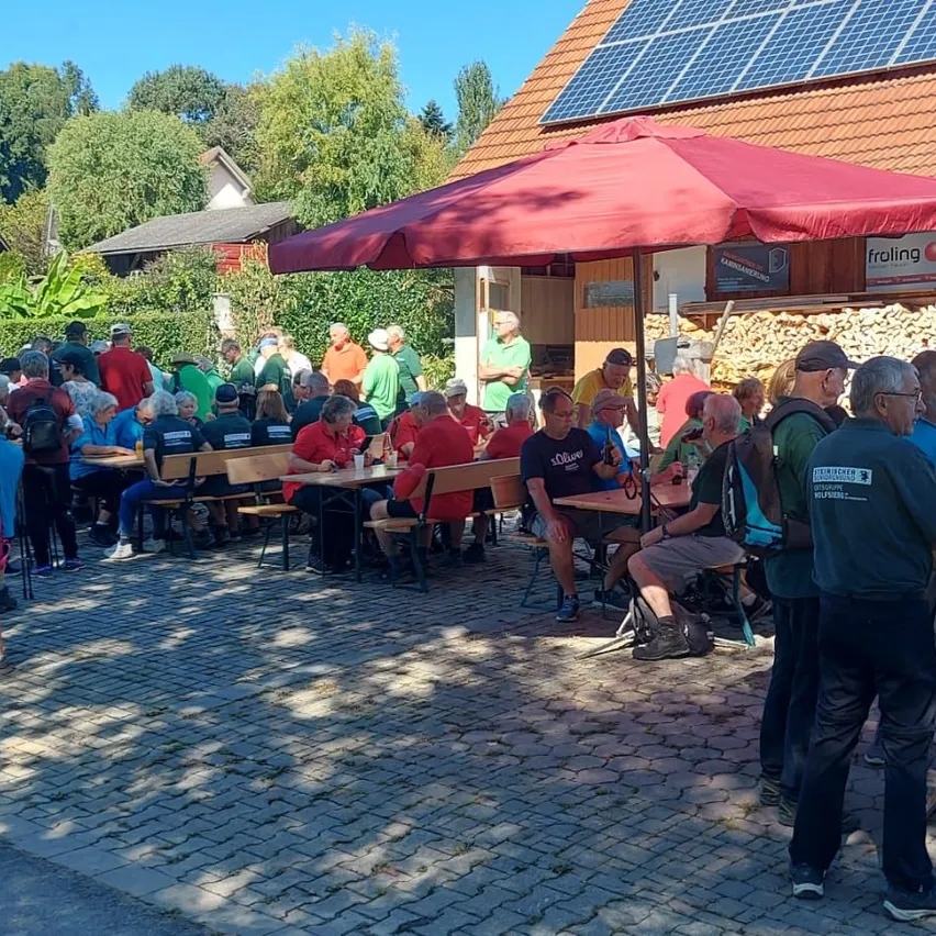 Eine Gruppe von Menschen sitzt unter einem Schirm um Tische herum vor einem Gebäude mit Solarzellen auf dem Dach.