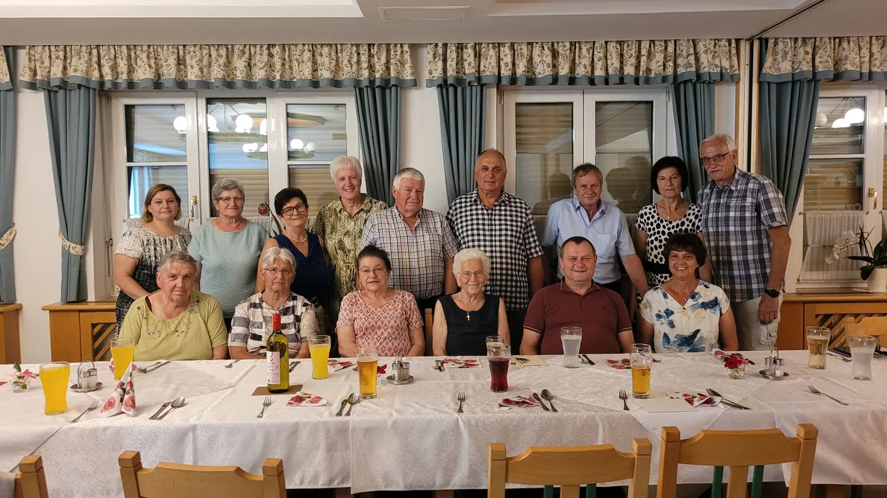 Eine Gruppe von Menschen versammelt sich um einen Esstisch mit Getränken und Essen. Sie alle lächeln und scheinen für ein Foto zu posieren. Der Tisch ist mit Tellern, Löffeln, Gabeln und Weinflaschen gedeckt. Der Raum hat große Fenster mit Vorhängen.