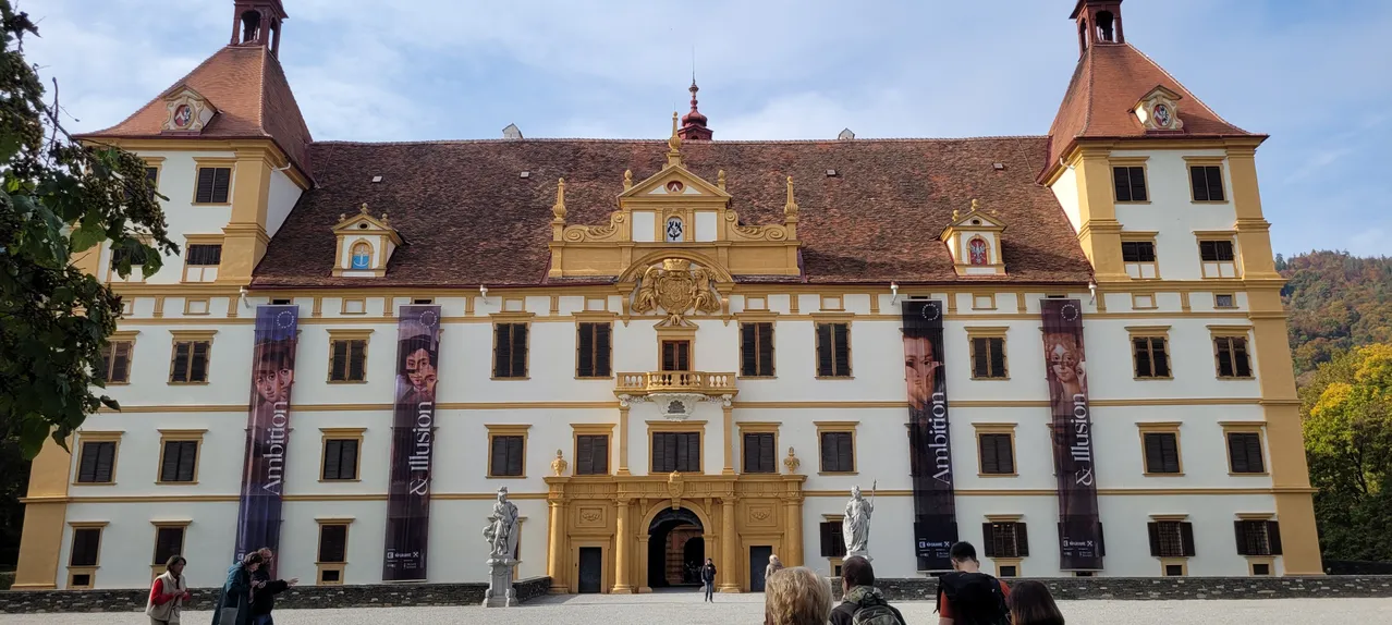 Ein golden akzentuiertes weißes Gebäude mit einem gefliesten Dach und vielen Fenstern steht vor einem blauen Himmel. Banner hängen an den Seiten. Zwei Statuen stehen auf beiden Seiten des Eingangs.