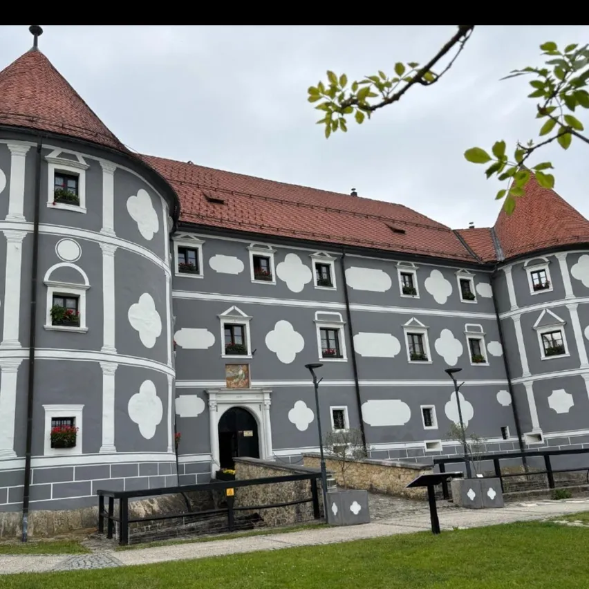 Ein historisches Schloss mit einem roten Dach und weißen gemusterten Wänden. Die Vorderseite zeigt eine schwarze Eingangstür, umgeben von Fenstern mit Topfpflanzen. Das Schloss ist von einem kleinen Garten und einem Metallzaun umgeben.