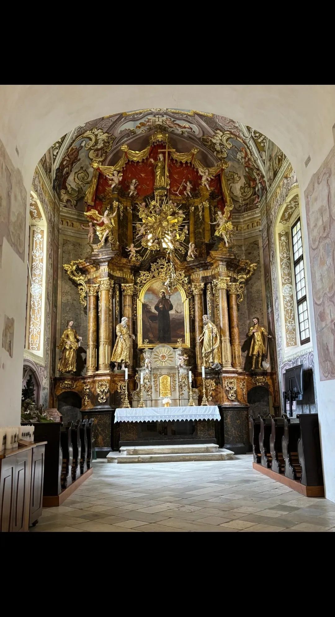 Ein barocker Kirchenaltar mit goldenen Statuen, einem zentralen Gemälde und aufwendigen Verzierungen. Zwei Kerzen flankieren den Altar. Bänke und eine Kanzel sind sichtbar.
