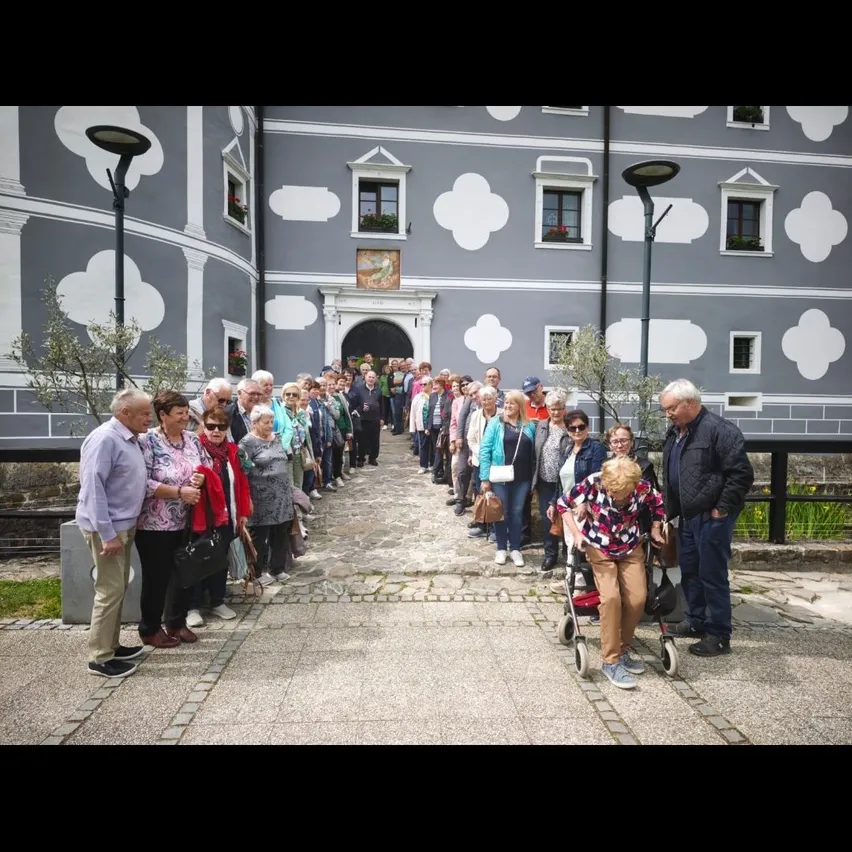 Eine Gruppe von Menschen, hauptsächlich ältere, warten in einer Schlange vor einem Gebäude mit dekorativen weißen Mustern. Einige tragen Handtaschen und einer schiebt einen Kinderwagen.
