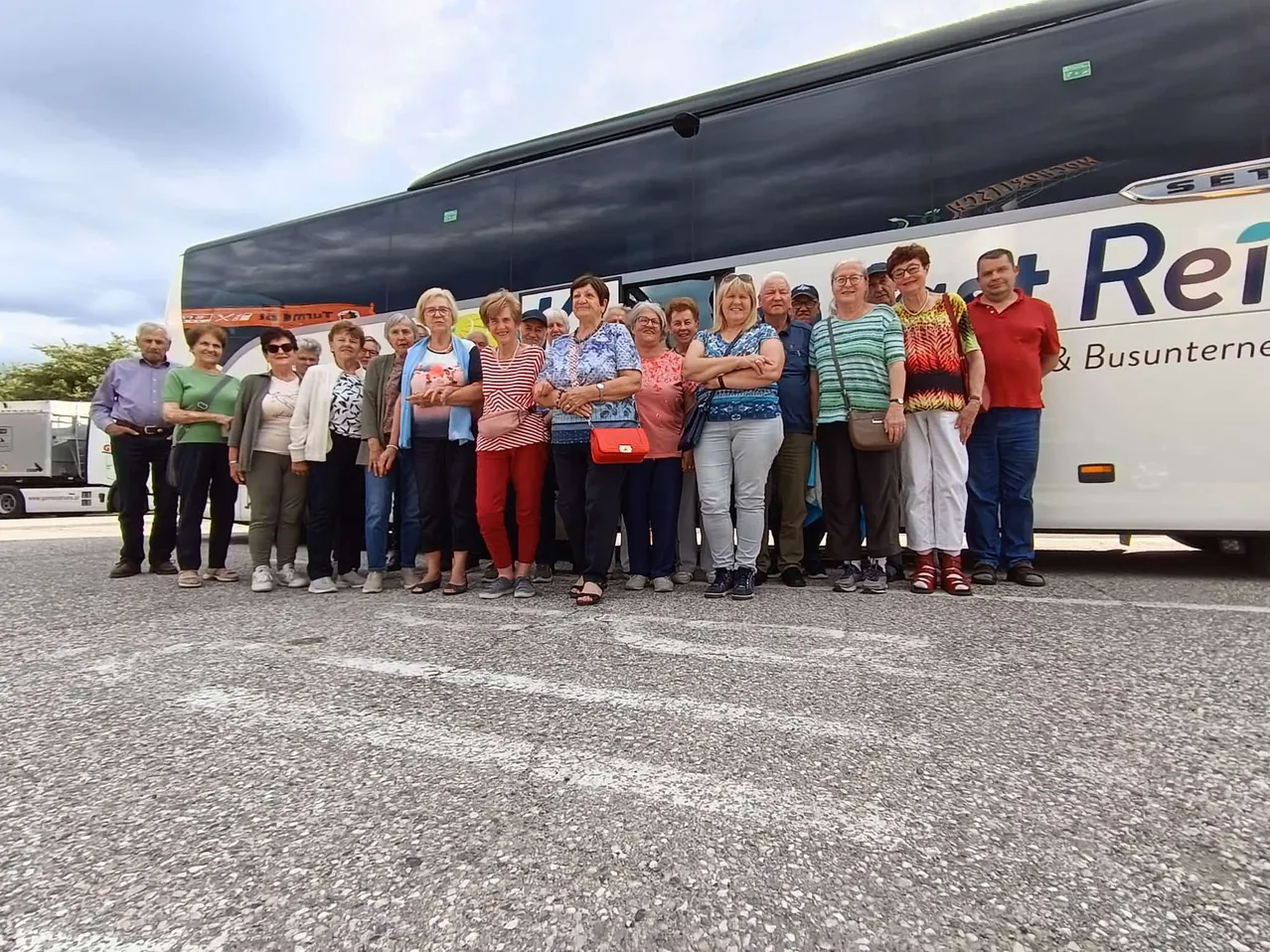 Eine Gruppe Erwachsener posiert vor einem Bus an einem sonnigen Tag. Sie tragen Freizeitkleidung, einige mit Brille. Der Bus hat weiße Schrift auf blauem Hintergrund.