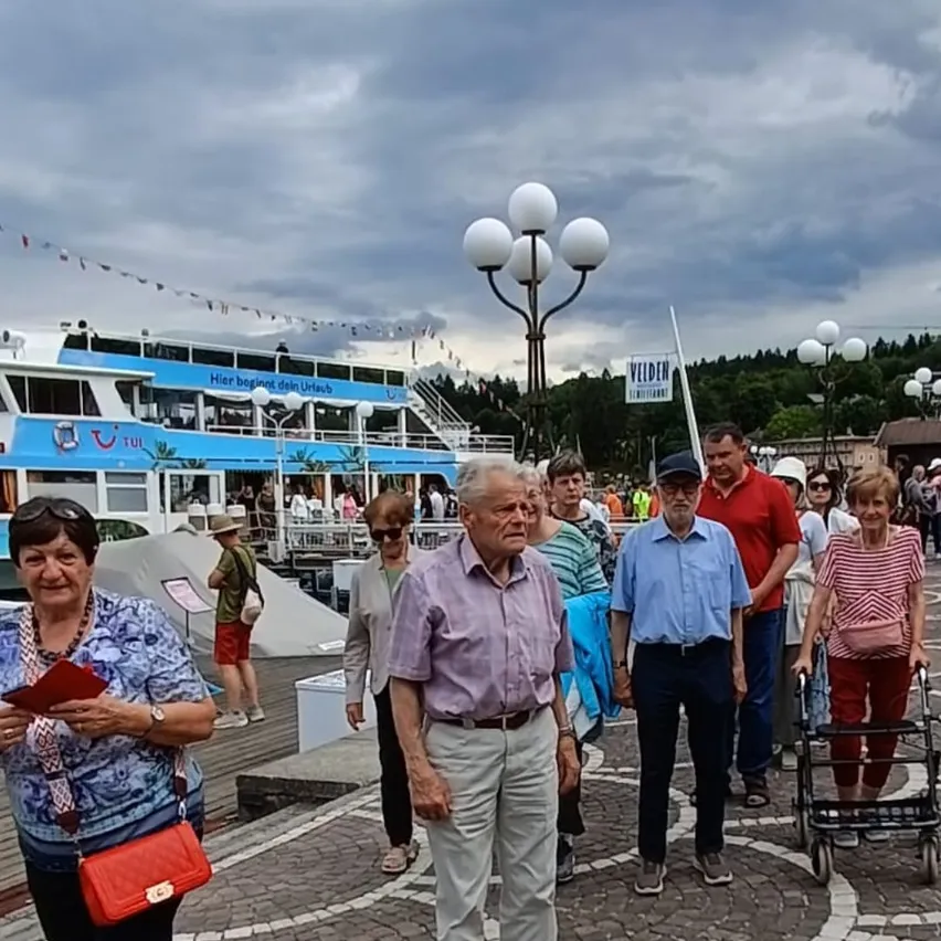 Eine Gruppe älterer Personen, einige mit Gehhilfen, gehen auf einem gepflasterten Steg. Ein Boot mit blau-weißem Farbschema und einem Schild, das 'Hier beginnt Ihr Urlaub' steht im Hintergrund.
