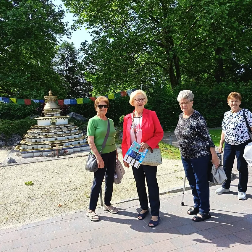 Vier Frauen stehen vor einem kleinen Steinstupa, einer dekorativen Pagode, in einem Park mit grünen Bäumen und bunten Flaggen. Sie tragen Freizeitkleidung und einige tragen eine Brille.
