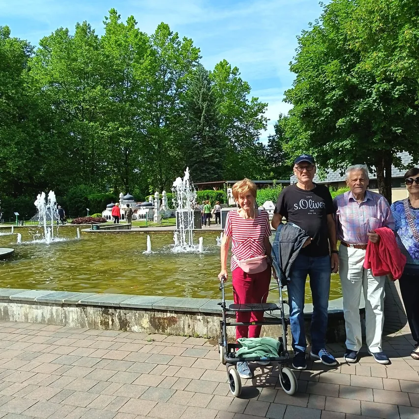 Eine Gruppe von Menschen steht an einem Brunnen in einem Park. Eine Frau in roten Hosen hält einen Gehstock. Hinter ihr stehen drei Leute. Der Mann auf der rechten Seite hält eine rote Jacke.