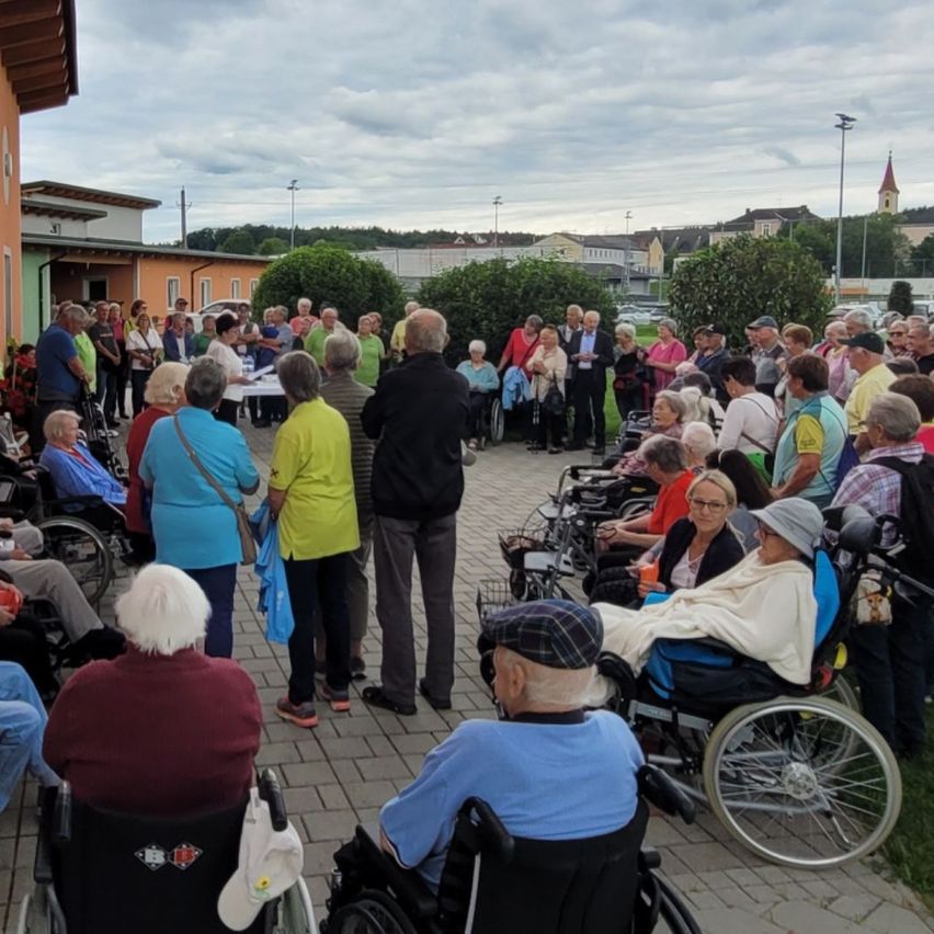 A large group of elderly people gathered outdoors, many seated in wheelchairs, with some standing. They appear to be attending an event, with a few people standing in front, possibly giving speeches. Trees and buildings are visible in the background.