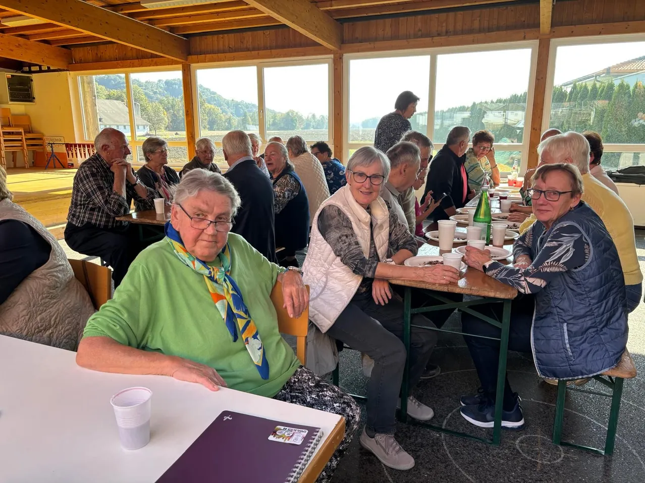 Eine Gruppe älterer Menschen sitzt um einen Tisch in einem Speisesaal. Sie tragen Brillen und einige tragen Schals. Der Tisch ist mit Tellern, Bechern und Flaschen gedeckt. Der Speisesaal hat große Fenster mit Blick nach draußen.
