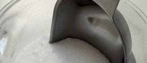 Eine Glasschale mit weißem Zucker, mit einem Metalllöffel, der darauf ruht. Die Schüssel steht auf einer weißen Oberfläche.