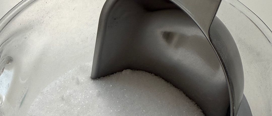 Eine Glasschale mit weißem Zucker, mit einem Metalllöffel, der darauf ruht. Die Schüssel steht auf einer weißen Oberfläche.