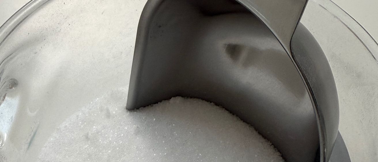Eine Glasschale mit weißem Zucker, mit einem Metalllöffel, der darauf ruht. Die Schüssel steht auf einer weißen Oberfläche.