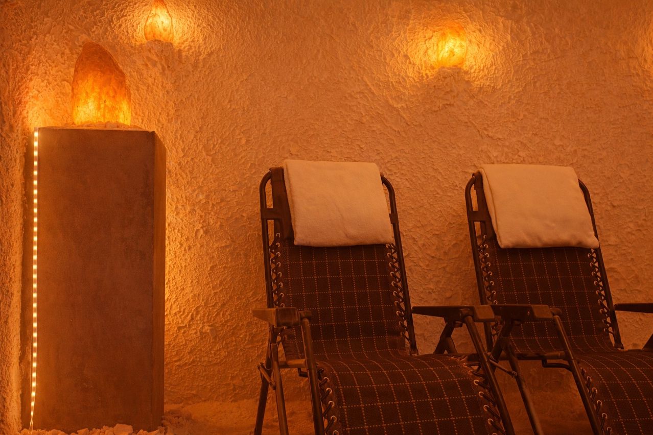 Zwei Liegestühle mit weißen Kissen befinden sich in einem schwach beleuchteten Raum mit gelber Wand und zwei Lampen. Der Boden ist mit kleinen Steinen bedeckt.
