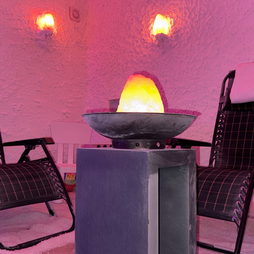 Ein kleiner Raum mit einem warmen rosa Schimmer zeigt einen zentralen beleuchteten Stein und zwei Liegestühle. Ein Stuhl hat ein weißes Kissen, und die Wände sind in Rosa gestrichen.