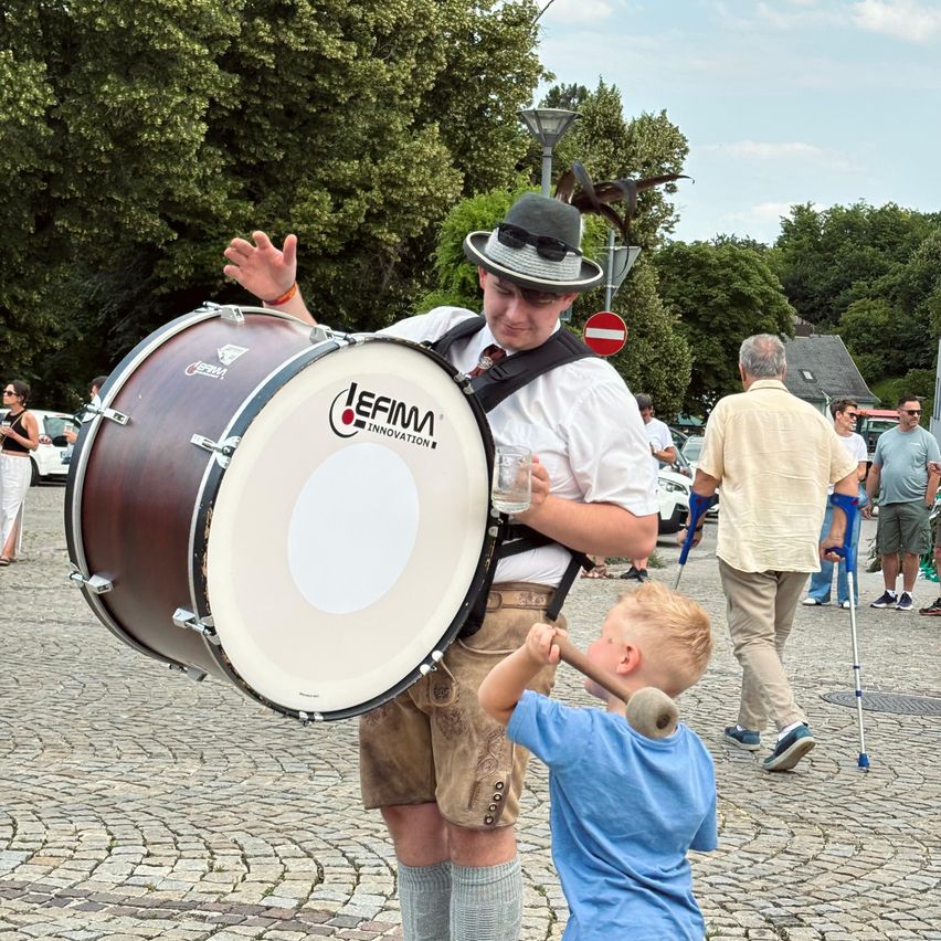 Ein Mann in traditioneller deutscher Kleidung trägt eine große Trommel, und ein Junge schlägt mit einem Hammer darauf. Im Hintergrund gehen Menschen.