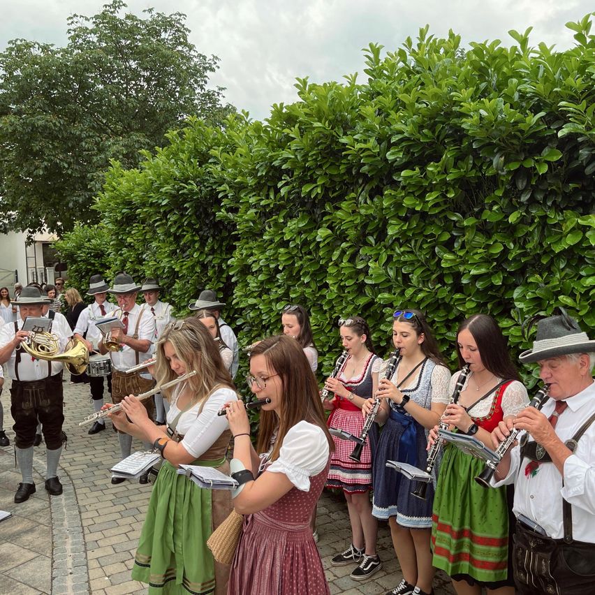 Eine Gruppe von Musikern in traditioneller Kleidung, die verschiedene Instrumente im Freien spielen. Dahinter bildet ein Busch einen Hintergrund, und einige Leute stehen weiter hinten.